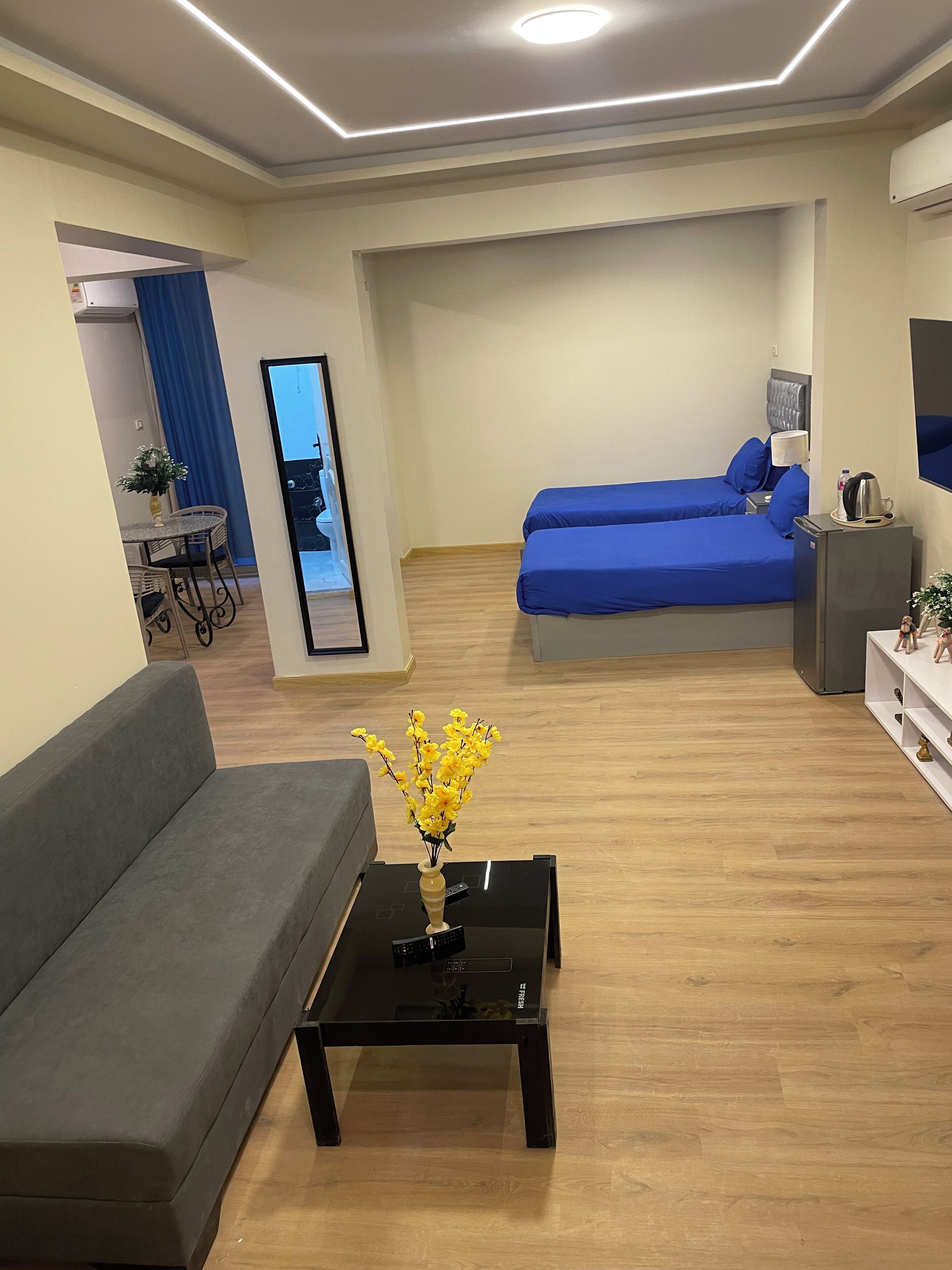 A modern living room with a grey sofa, a black coffee table with a vase of yellow flowers, and a bed with blue bedding.