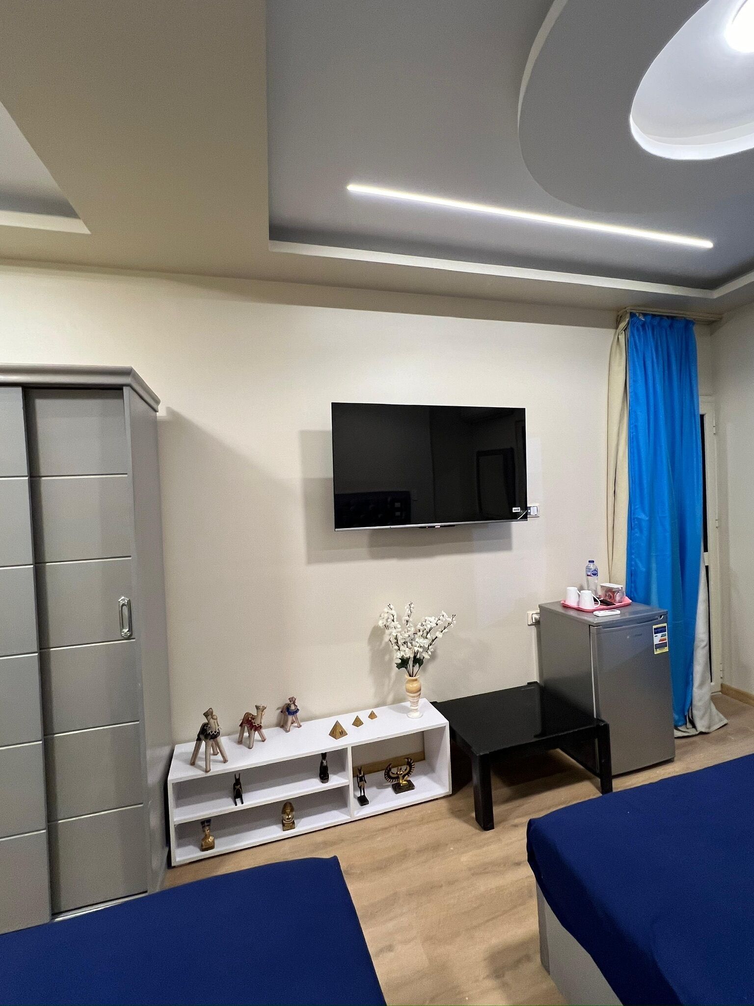 A modern hotel room with a grey wardrobe, a flat-screen TV mounted on the wall, a small white table with decorative items, a blue curtain, and a bed with a blue bedspread.