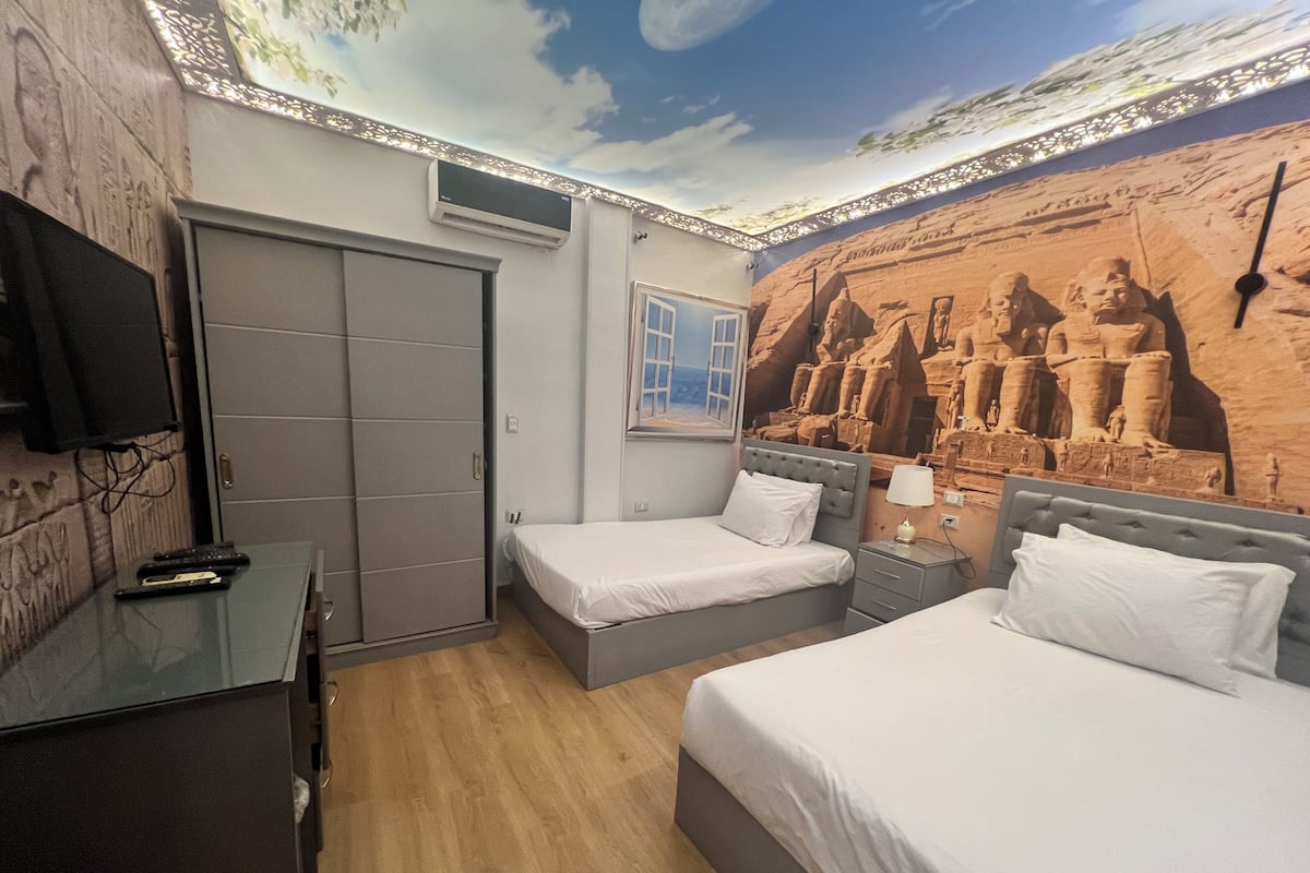 A hotel room with two beds, a TV, a wardrobe, and a mural of ancient Egyptian figures.