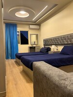 A hotel room with two beds, a large mirror, a tufted headboard, a bedside table with a lamp, and a window with blue curtains.