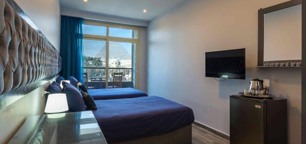 Habitació d'hotel amb un llit, una televisió, escriptori amb làmpada i finestra amb vista a una piràmide.