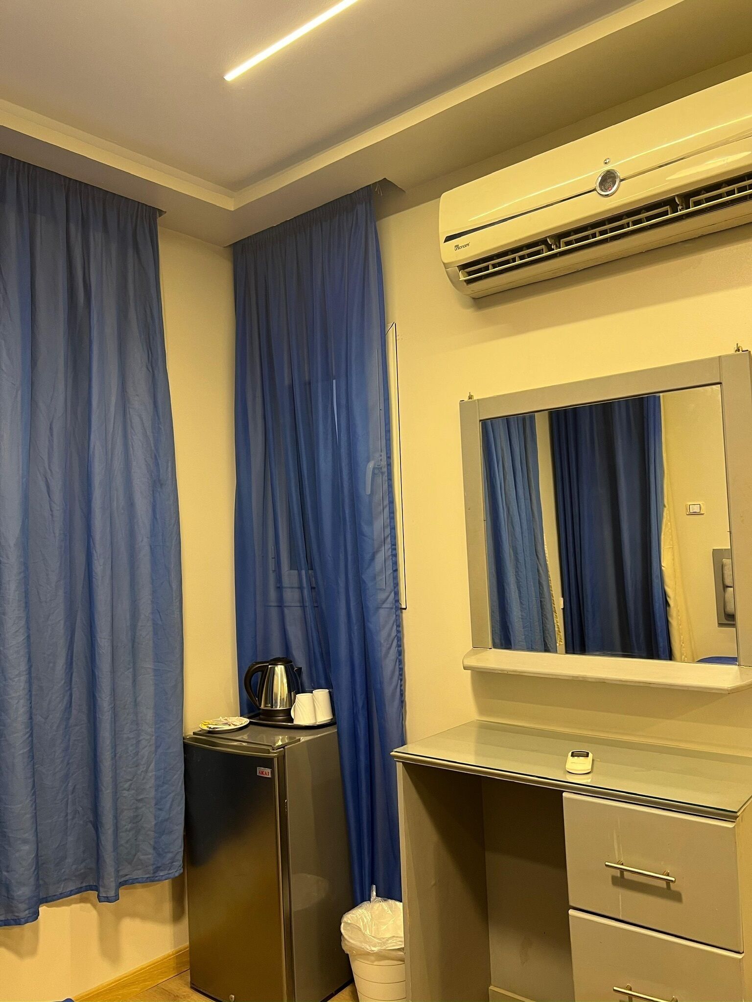 A hotel room with a minibar, a refrigerator, a trash bin, a desk with drawers, a mirror, and blue curtains.