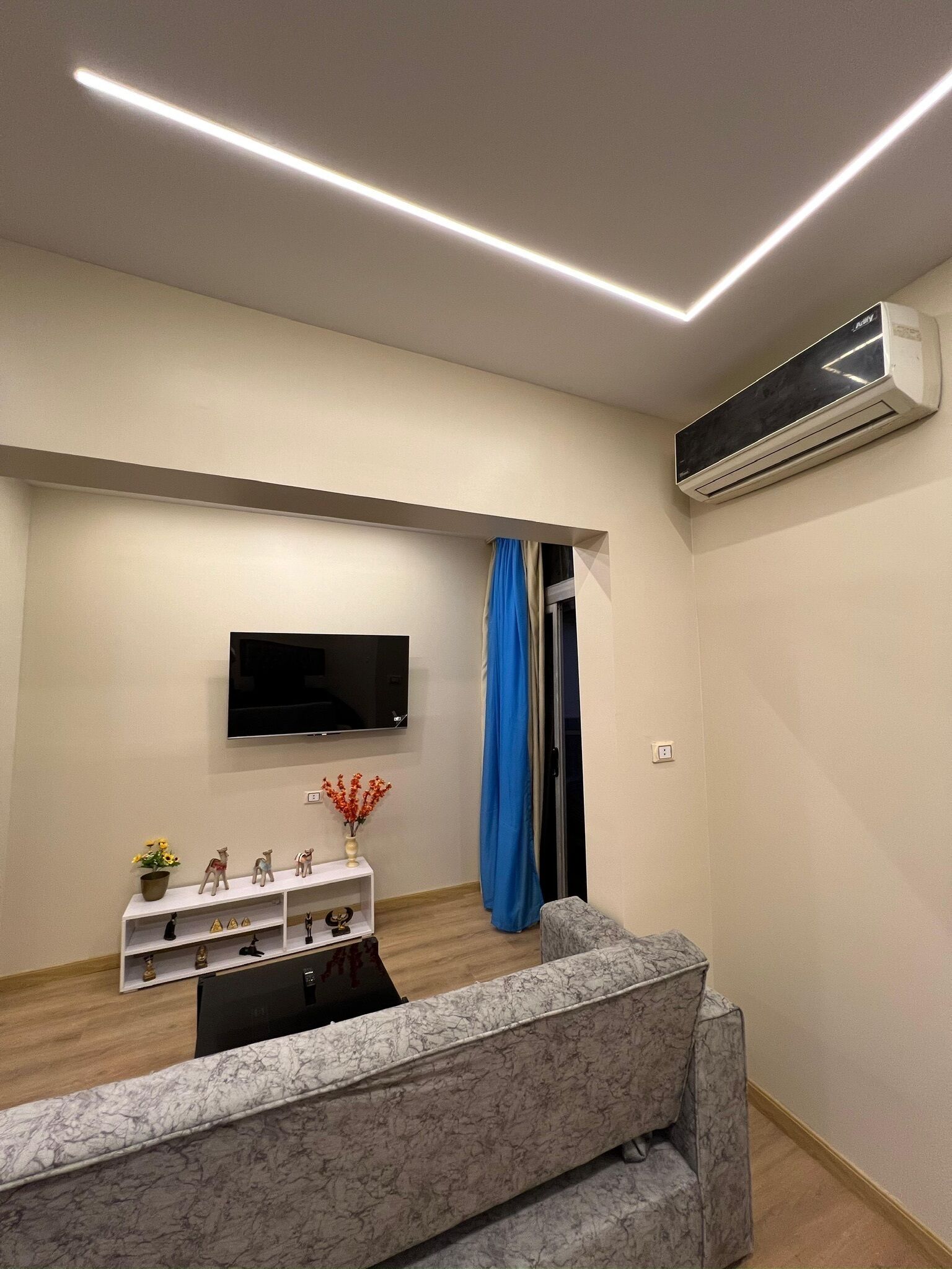 Een moderne woonkamer met een flatscreen-tv, een bank met marmerlook, een witte plank met decoratieve objecten, en een airconditioning.