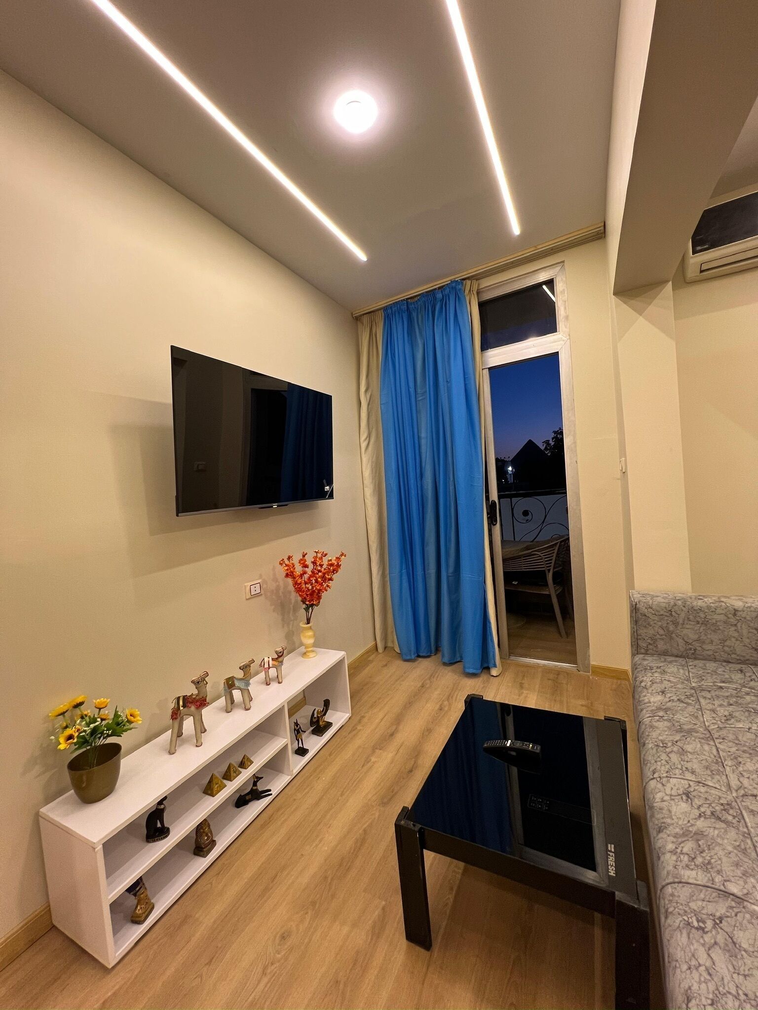 Una sala de estar moderna con una televisión de pantalla plana, una mesa de centro y un sofá. Hay un estante blanco con adornos y una ventana con cortinas azules.