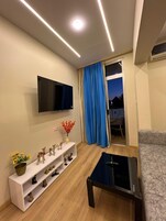 Una sala de estar moderna con una televisión de pantalla plana, una mesa de centro y un sofá. Hay un estante blanco con adornos y una ventana con cortinas azules.