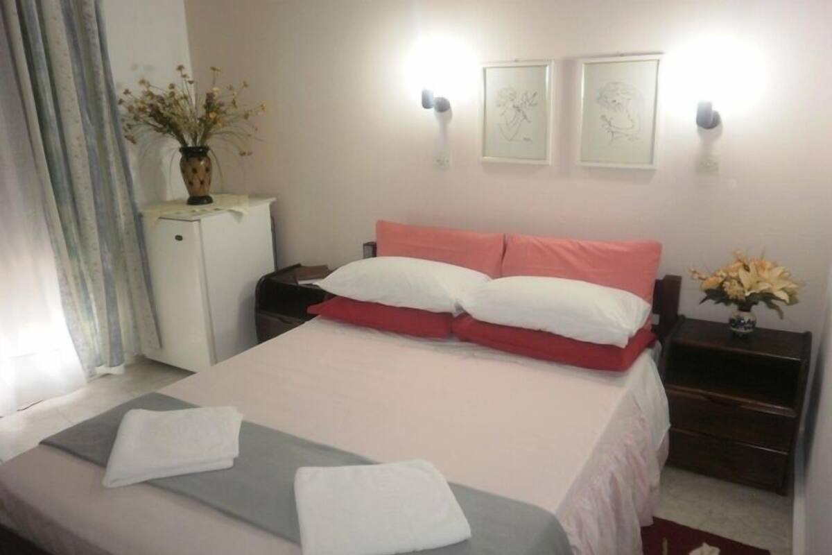 A neatly made bed with white and pink bedding, a small bedside table with a vase of flowers, and two framed artworks on the wall.