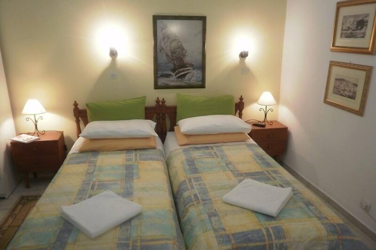 A double bed with a plaid bedspread, two pillows, and a folded towel on each bed. There are two bedside tables with lamps, a wall-mounted painting, and framed pictures on the wall.