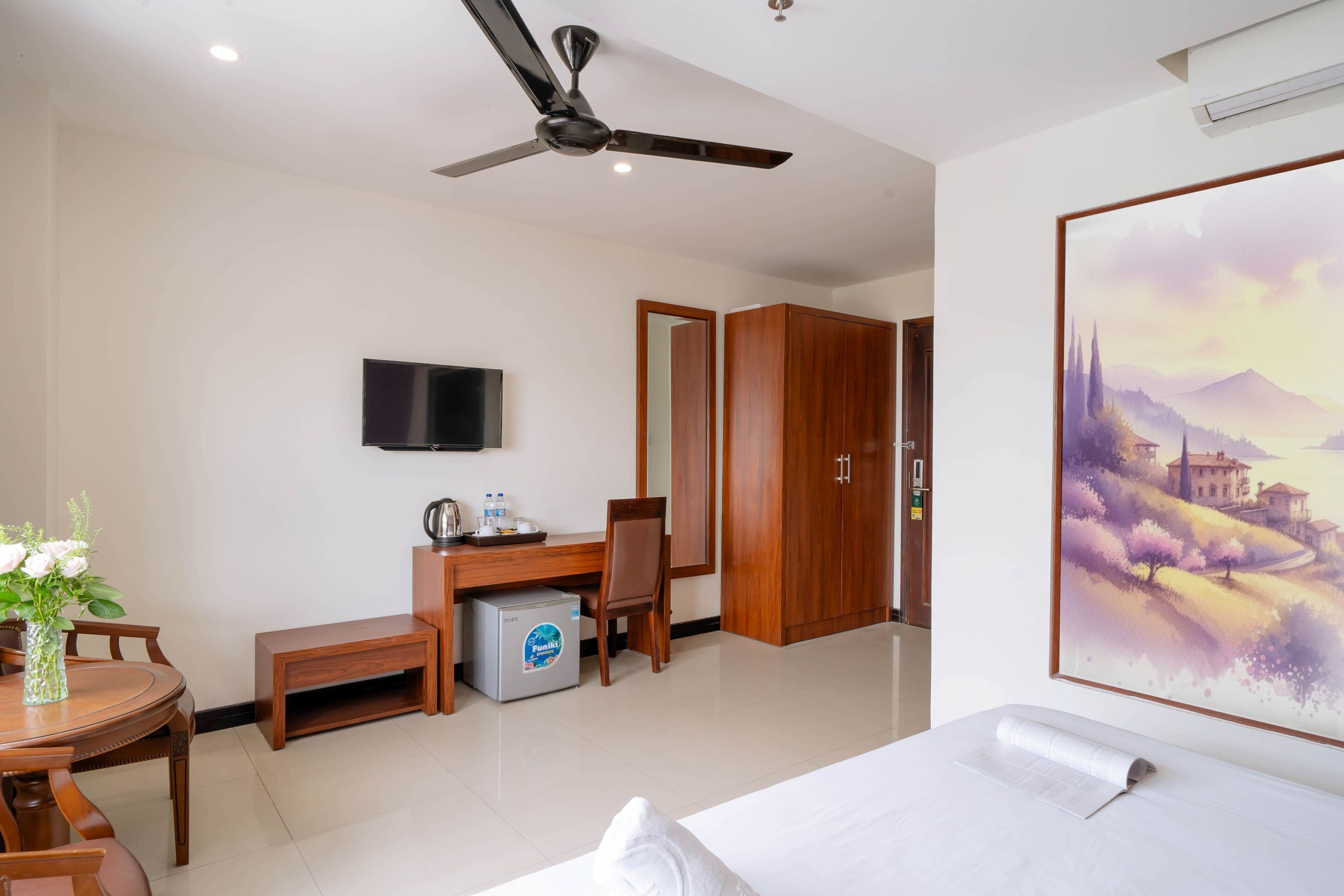 Une chambre d’hôtel avec un lit, un bureau et une chaise, un petit réfrigérateur, un téléviseur fixé au mur et un tableau accroché au mur.