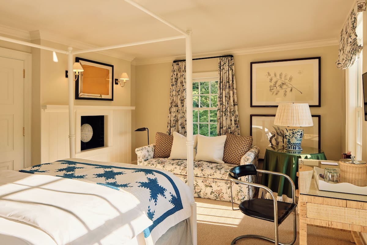 A bedroom with a four-poster bed, a green upholstered sofa, a wicker side table, and a framed botanical print on the wall.