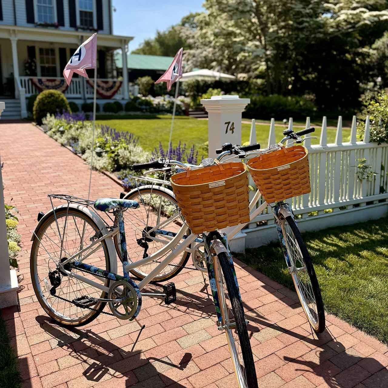 A bicycle with baskets parked on a brick pathway in front of a house with a white picket fence and a mailbox marked with the number 74.