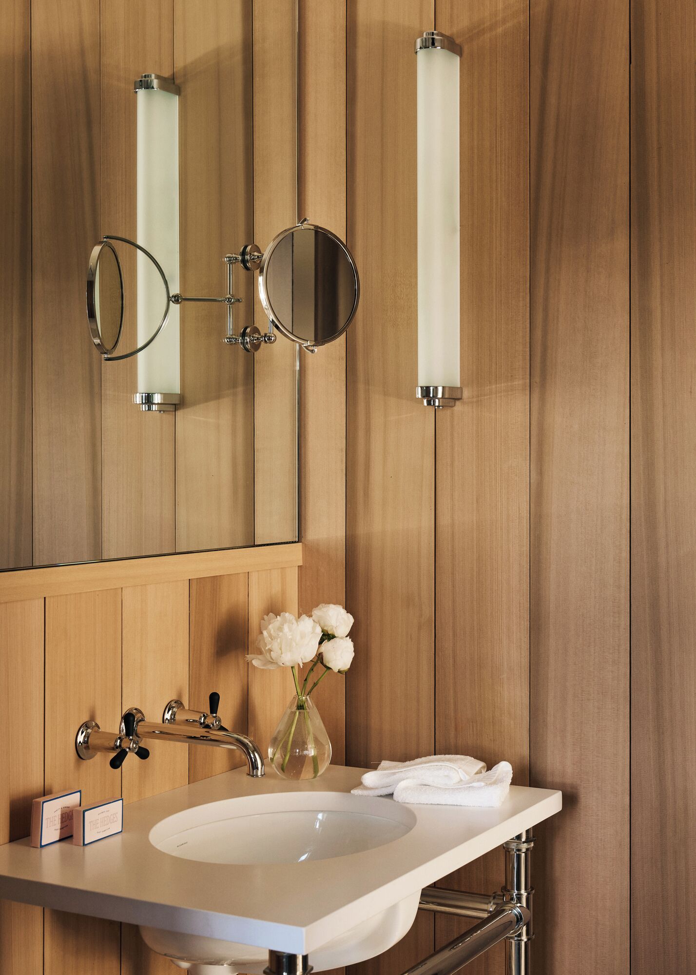 A bathroom with a wooden panel wall, a white sink, two round mirrors, and a vase with flowers.