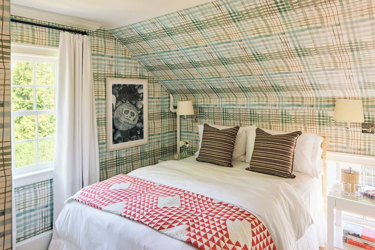 A bedroom with a checkered ceiling, a bed with pillows, a framed picture, and a window with curtains.