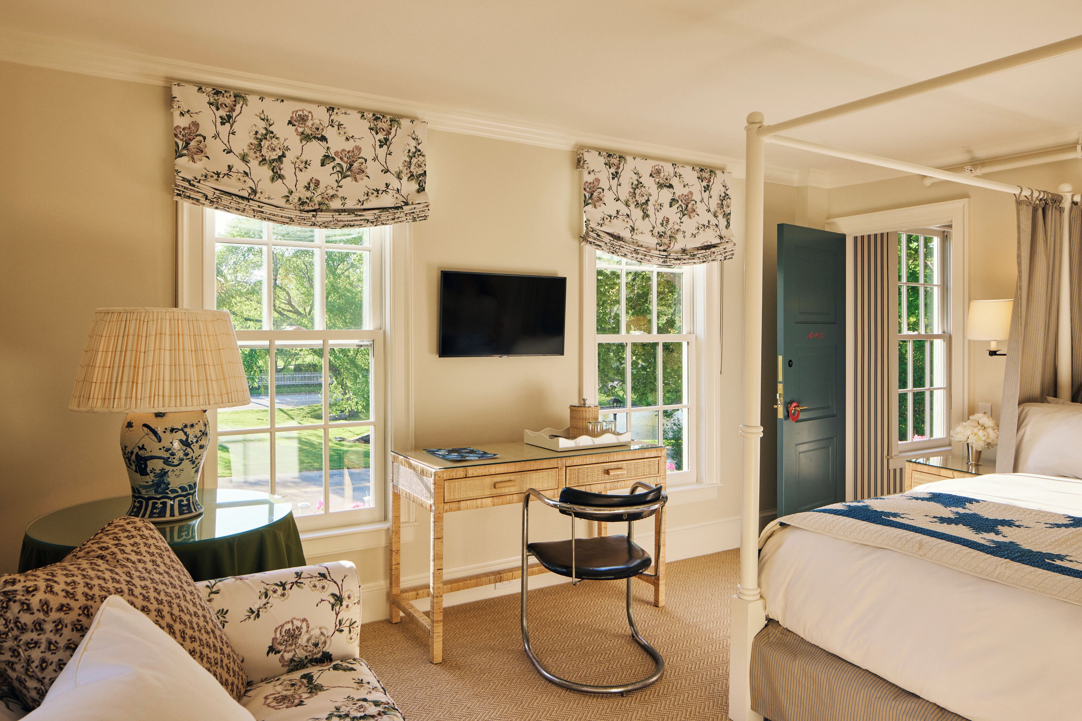 A bedroom with a bed, a desk, a chair, a television, and large windows with curtains.