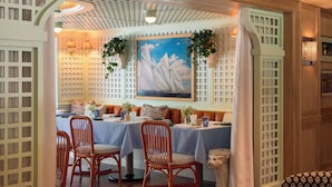Un coin repas avec une table dressée pour deux personnes, des chaises en osier, un tableau représentant des voiliers et un plafond en bois avec un motif quadrillé.