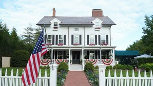 Une maison blanche à deux étages, avec un chemin de briques menant à la porte d’entrée, entourée d’une clôture en bois peinte en blanc et d’un drapeau.