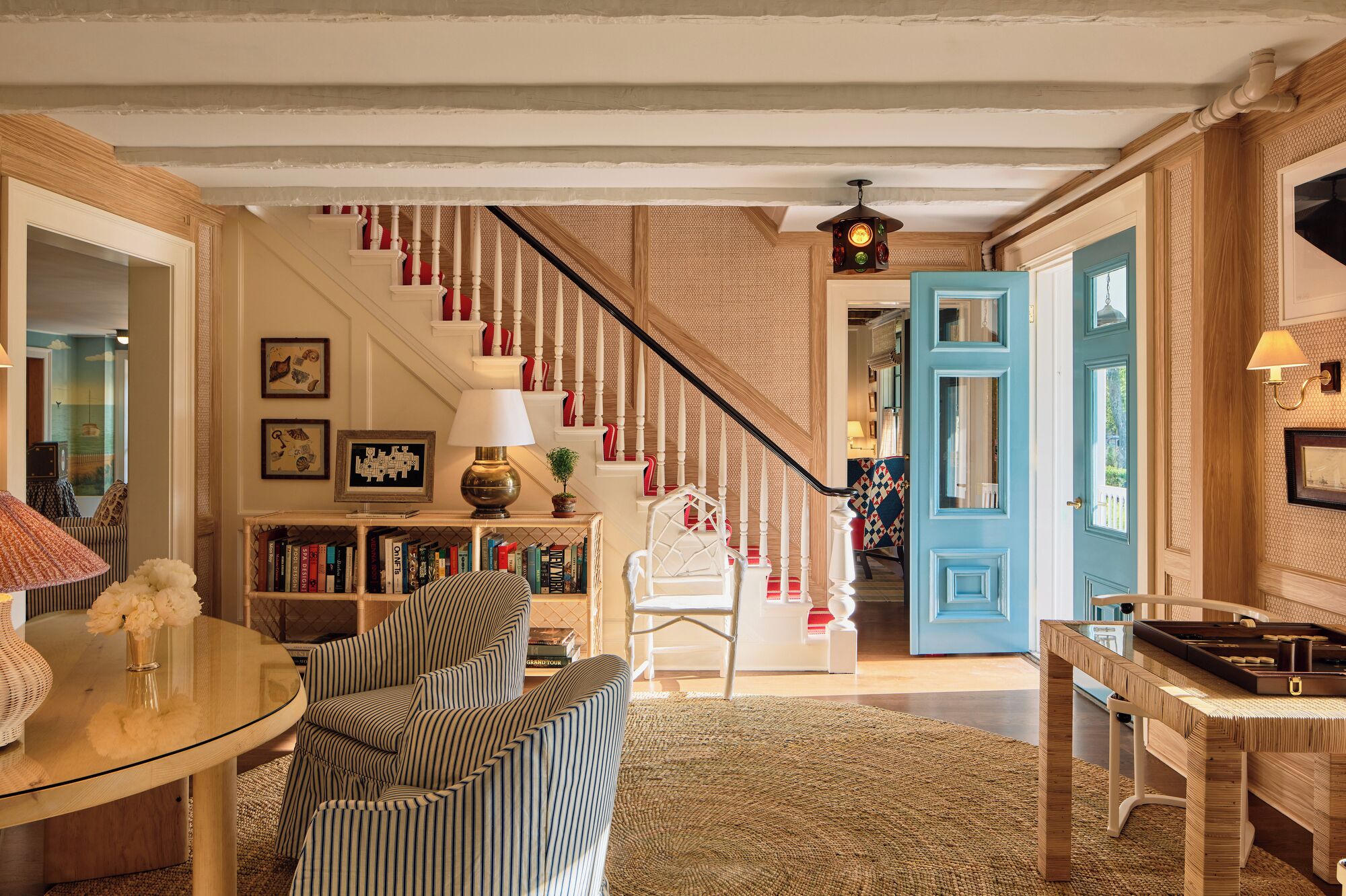 A living room with a staircase, a coffee table, striped chairs, a bookshelf, and a blue door.