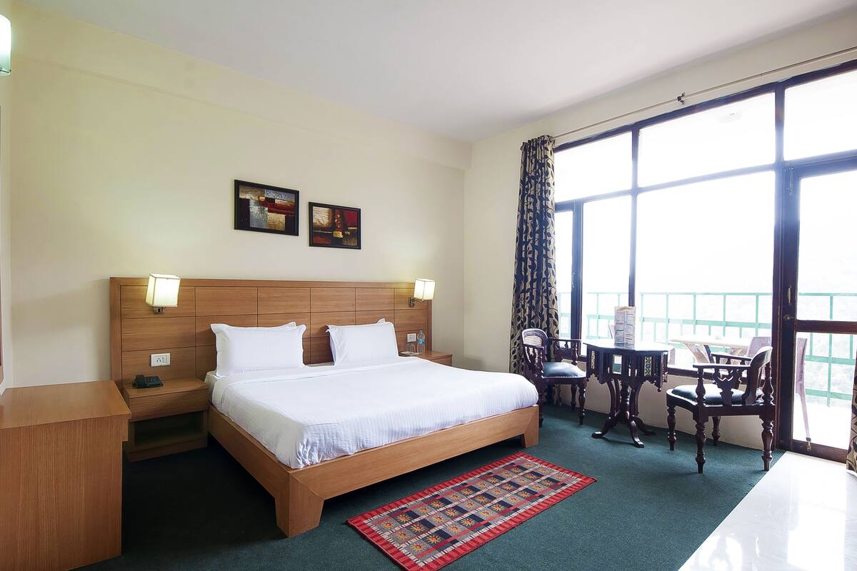 A hotel room with a large bed, bedside lamps, a small table with chairs, and a view through large windows.