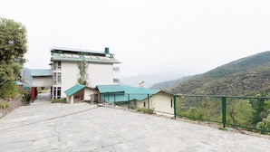 A building with a stone-paved entrance, a green-roofed structure, and a view of a hilly landscape. - The Ayali Fernhill Resort, Chail (Kandaghat)