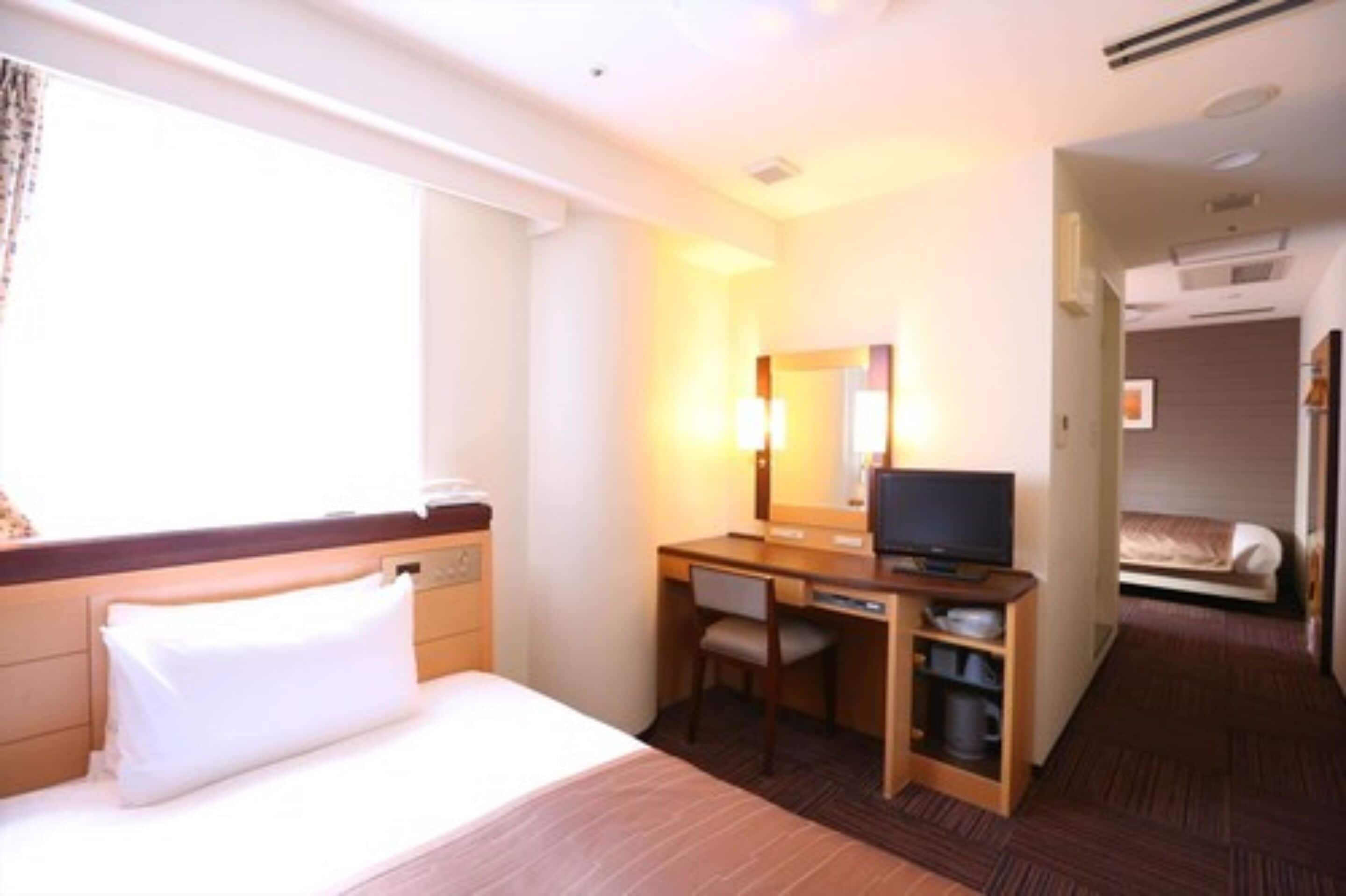 A hotel room with a bed, a desk with a chair, a television, and a small kitchenette.