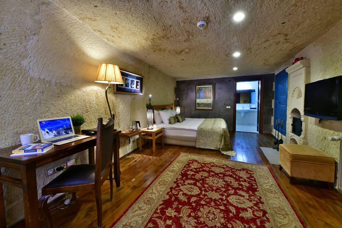 A cozy room with a wooden desk, a laptop, a bed, a TV, and a red patterned rug.