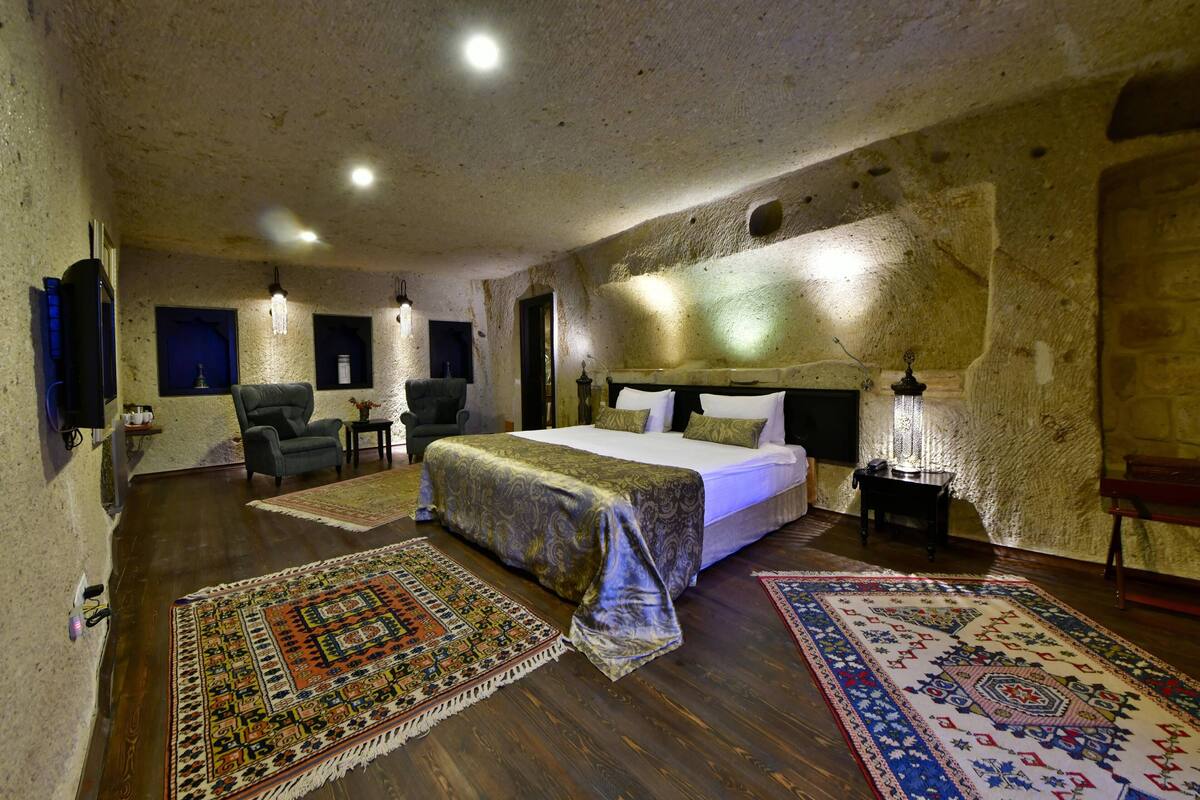 A spacious bedroom with a large bed, two armchairs, a small table, and a nightstand. The room features exposed stone walls and wooden flooring.