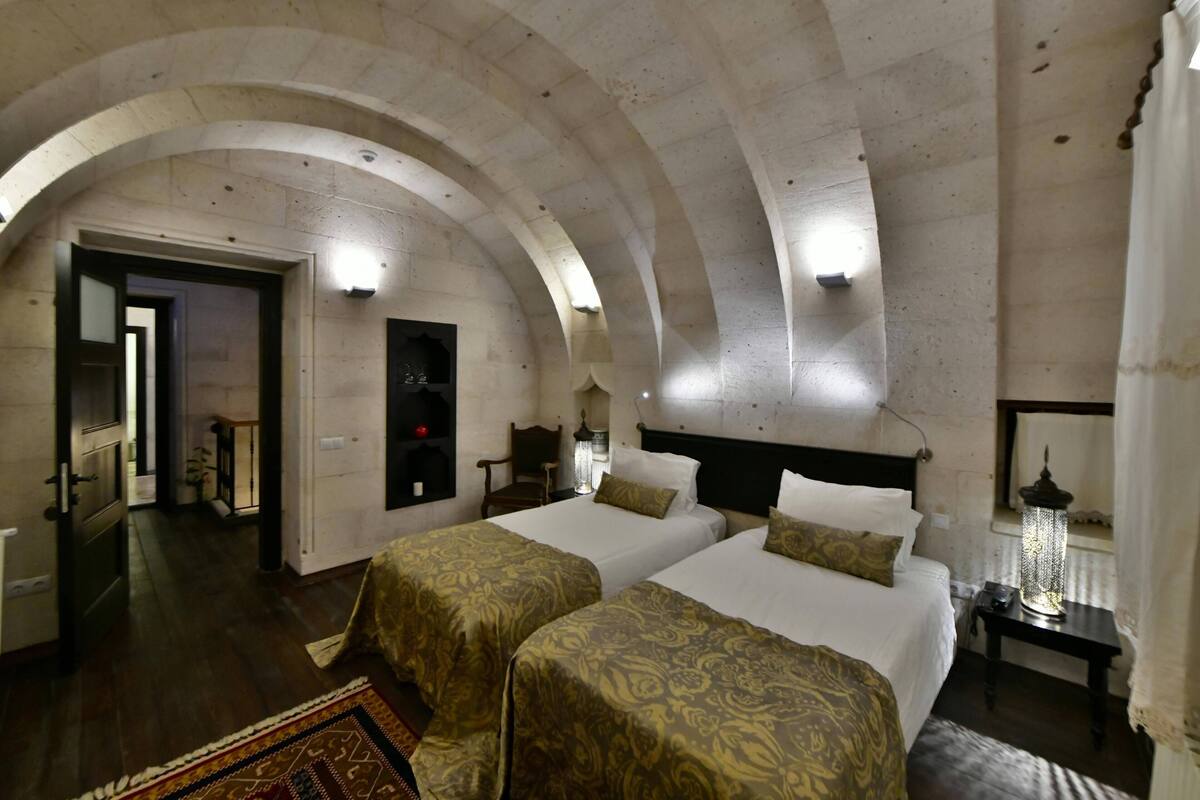 A room with two beds, a wooden floor, a chandelier, and a view through an arched doorway.