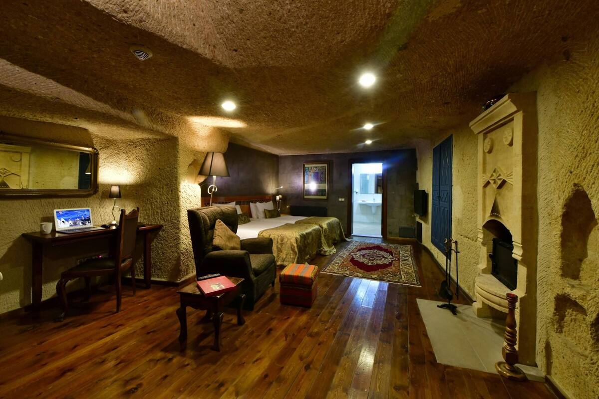 A rustic room with a wooden floor, a bed with a patterned bedspread, a desk with a laptop, a chair, and a small table with a book.