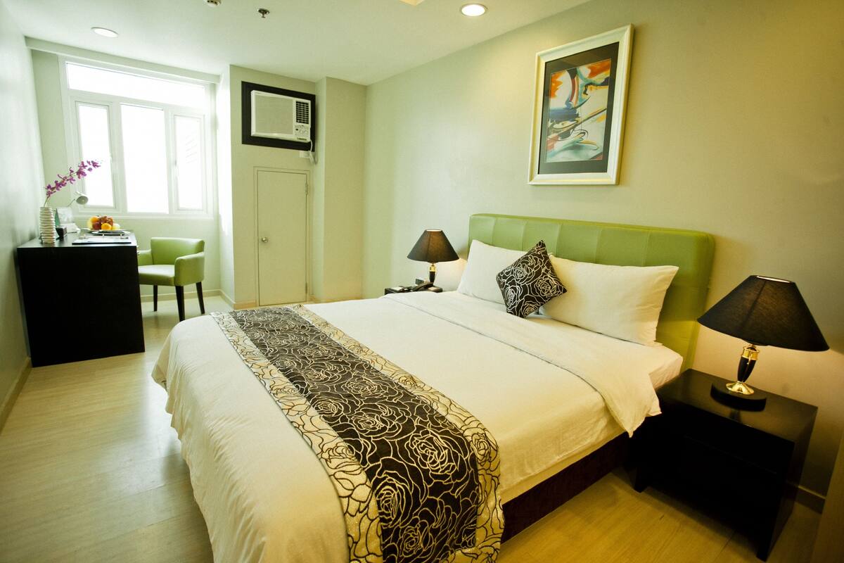 A hotel room with a large bed, a desk with a chair, a nightstand with a lamp, and a painting on the wall.
