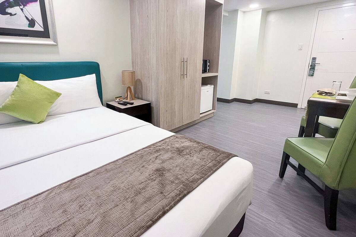 A hotel room with a bed, a green chair, a wardrobe, and a desk with a lamp.