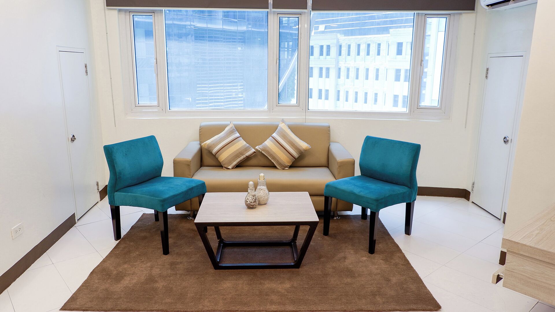 A room with a beige sofa, two teal armchairs, a coffee table, and a view of buildings through large windows.
