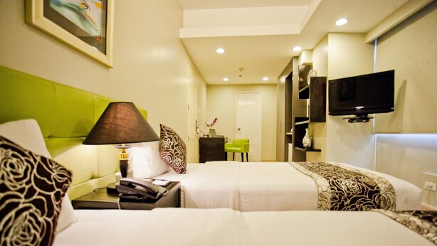 A hotel room with two beds, a desk with a lamp, a television, and a green chair. - The Exchange Regency Residence Hotel (Pasig)