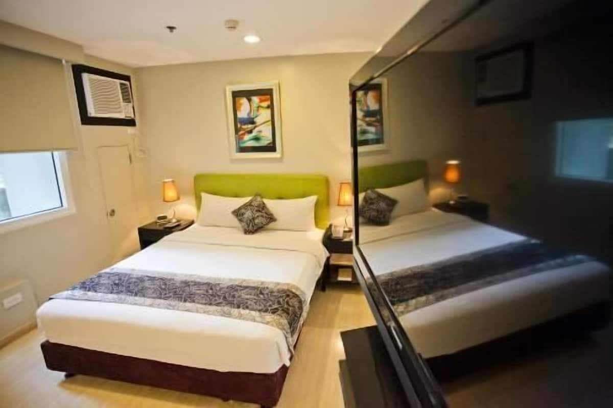 A double bed with a green headboard, two bedside tables with lamps, a window with blinds, and a mirror reflecting the room.