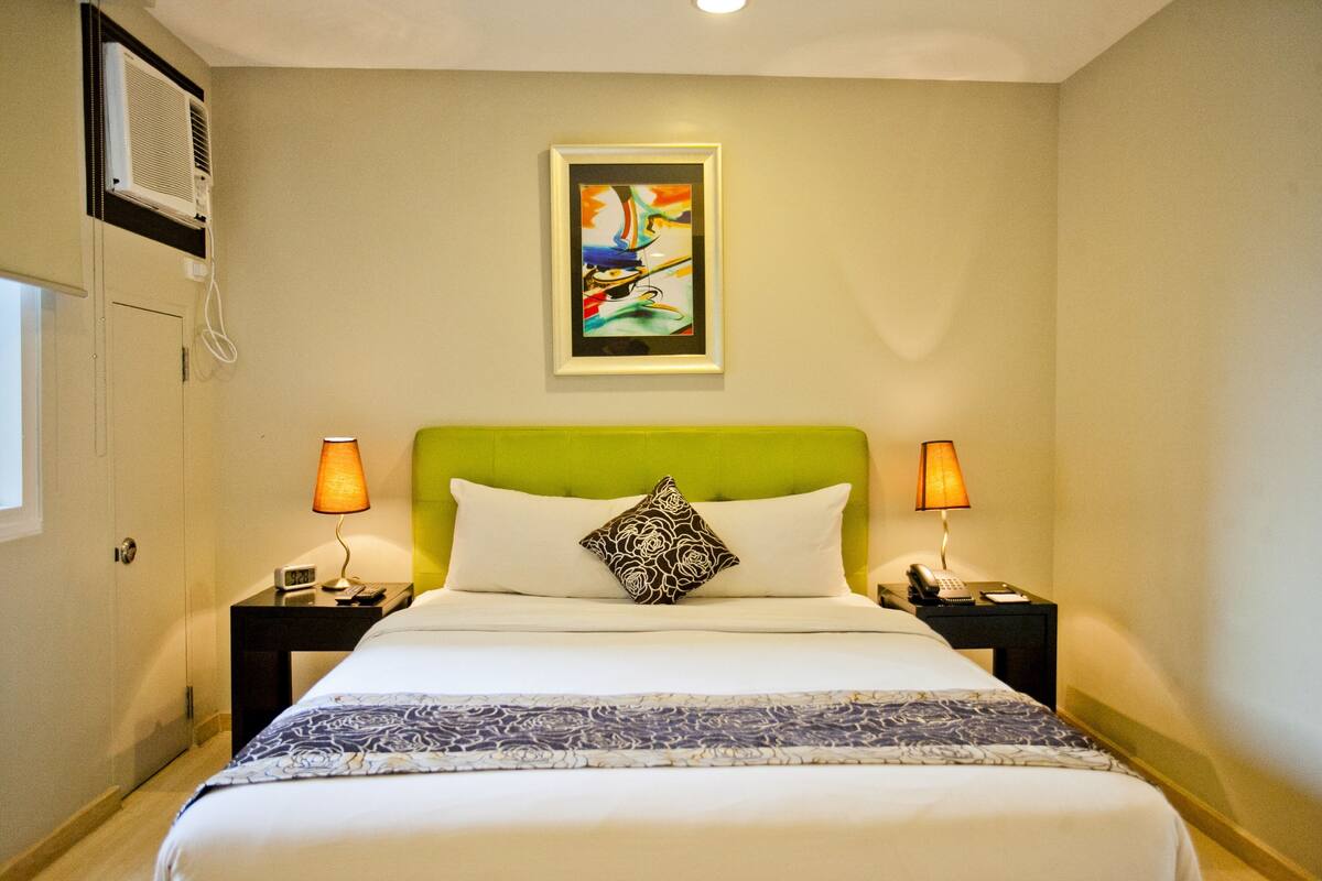A hotel room with a green headboard, white bedding, a patterned throw, two bedside lamps, a framed artwork on the wall, and a window on the left.