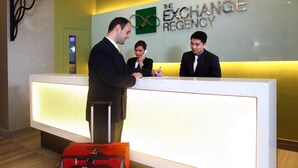 A reception desk at The Exchange Regency hotel with a modern design, a logo on the wall, and a red suitcase in front. - The Exchange Regency Residence Hotel (Pasig)