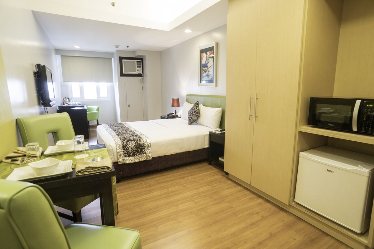 A hotel room with a bed, a desk with chairs, a microwave, and a small refrigerator.