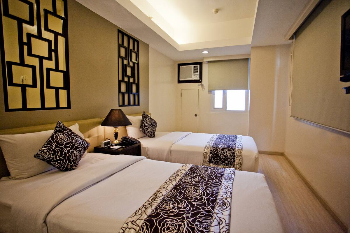 A hotel room with two beds, a nightstand with a lamp, a wall-mounted air conditioner, and a window with blinds.
