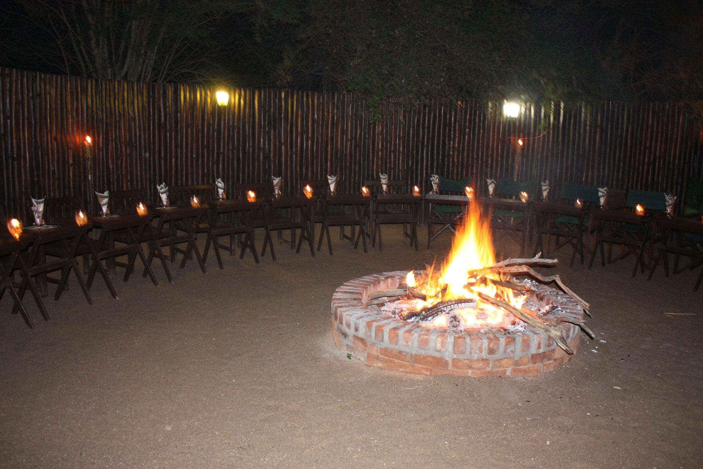 A circular fire pit with flames, surrounded by wooden chairs and tables, set against a bamboo fence.