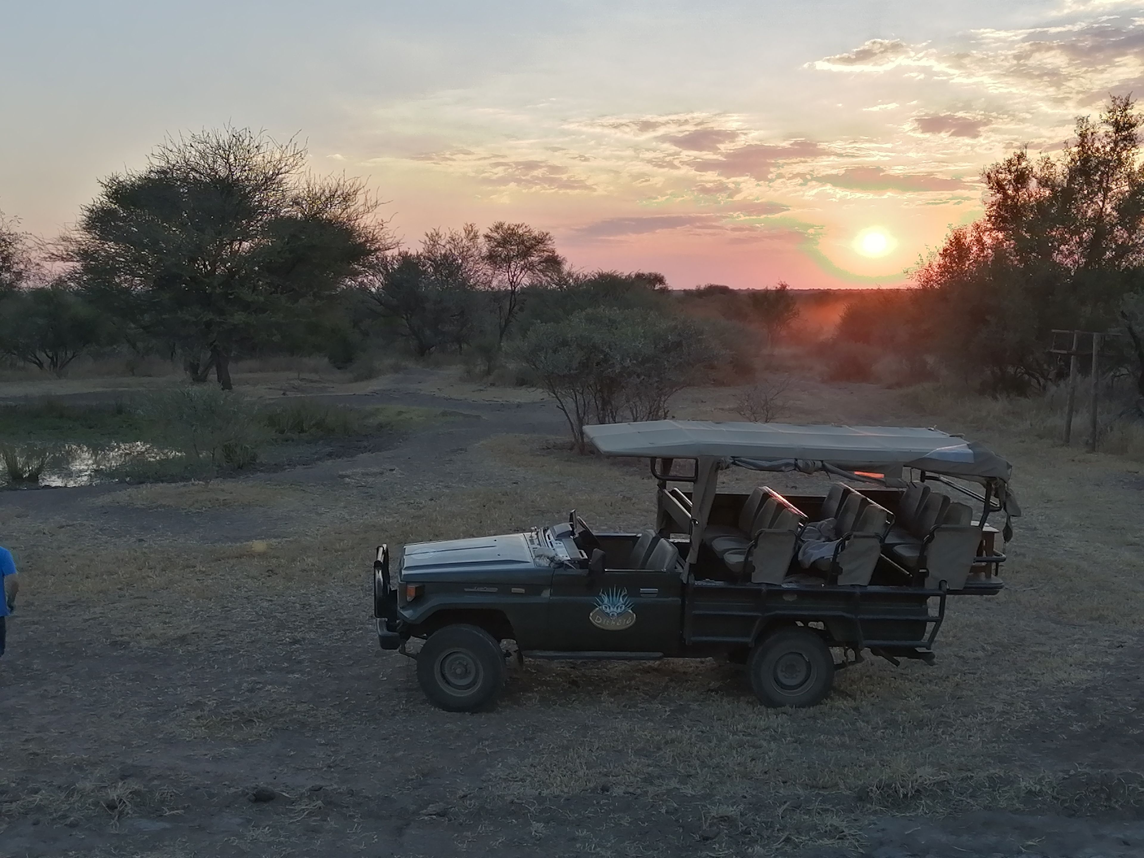 A safari vehicle parked in a dry, open landscape with sparse vegetation and a sunset in the background.