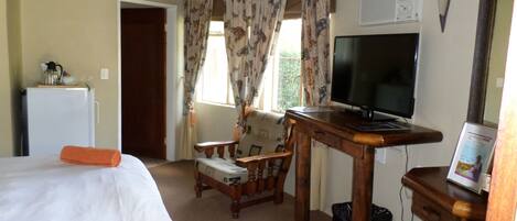 A hotel room with a bed, a desk with a TV, a chair, a small table, and a refrigerator.
