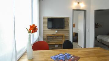 Ein modernes Zimmer mit Holztisch, Blumenvase, Wandfernseher und einer Couch im Hintergrund.