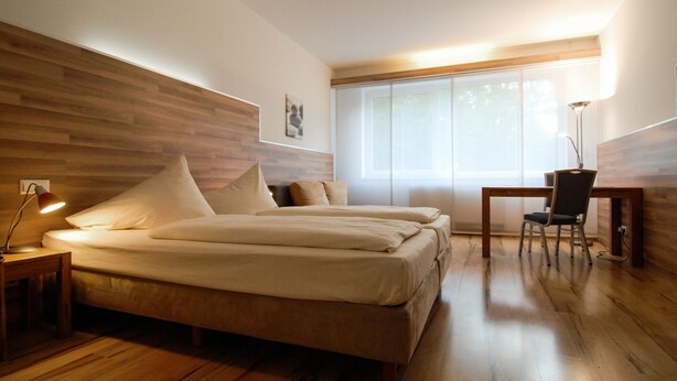 Un dormitorio con cabecera de madera, cama con ropa blanca, mesita de noche con lámpara, escritorio con silla y ventana con persianas.