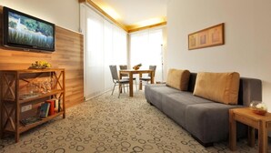 Una sala de estar moderna con un sofá gris, una mesa de centro de madera y un televisor montado en la pared. Hay una zona de comedor con una mesa y sillas, y una estantería llena de libros y vasos.