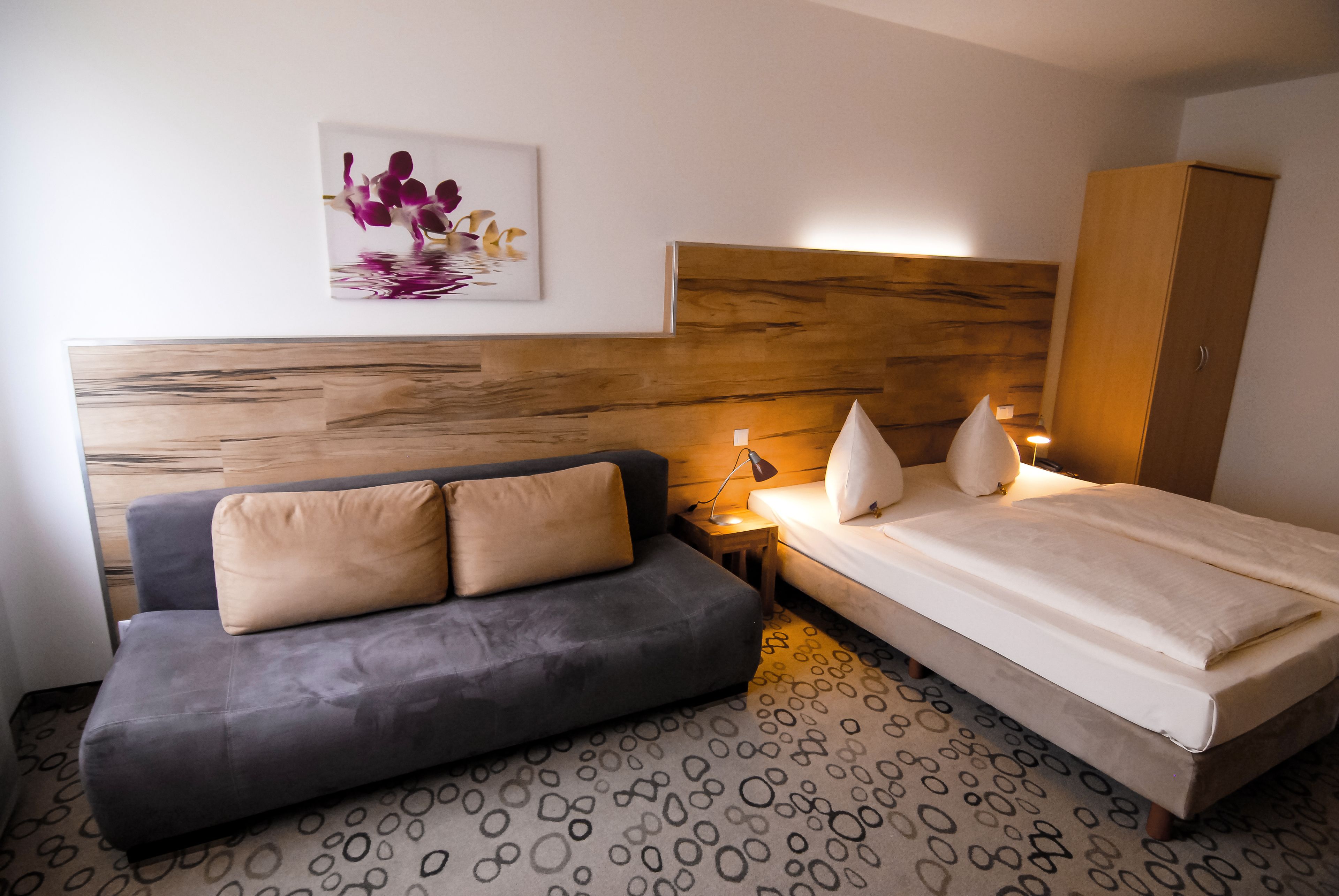 A bedroom with a wooden headboard, a grey sofa, a bed with white bedding, and a small bedside table with a lamp.