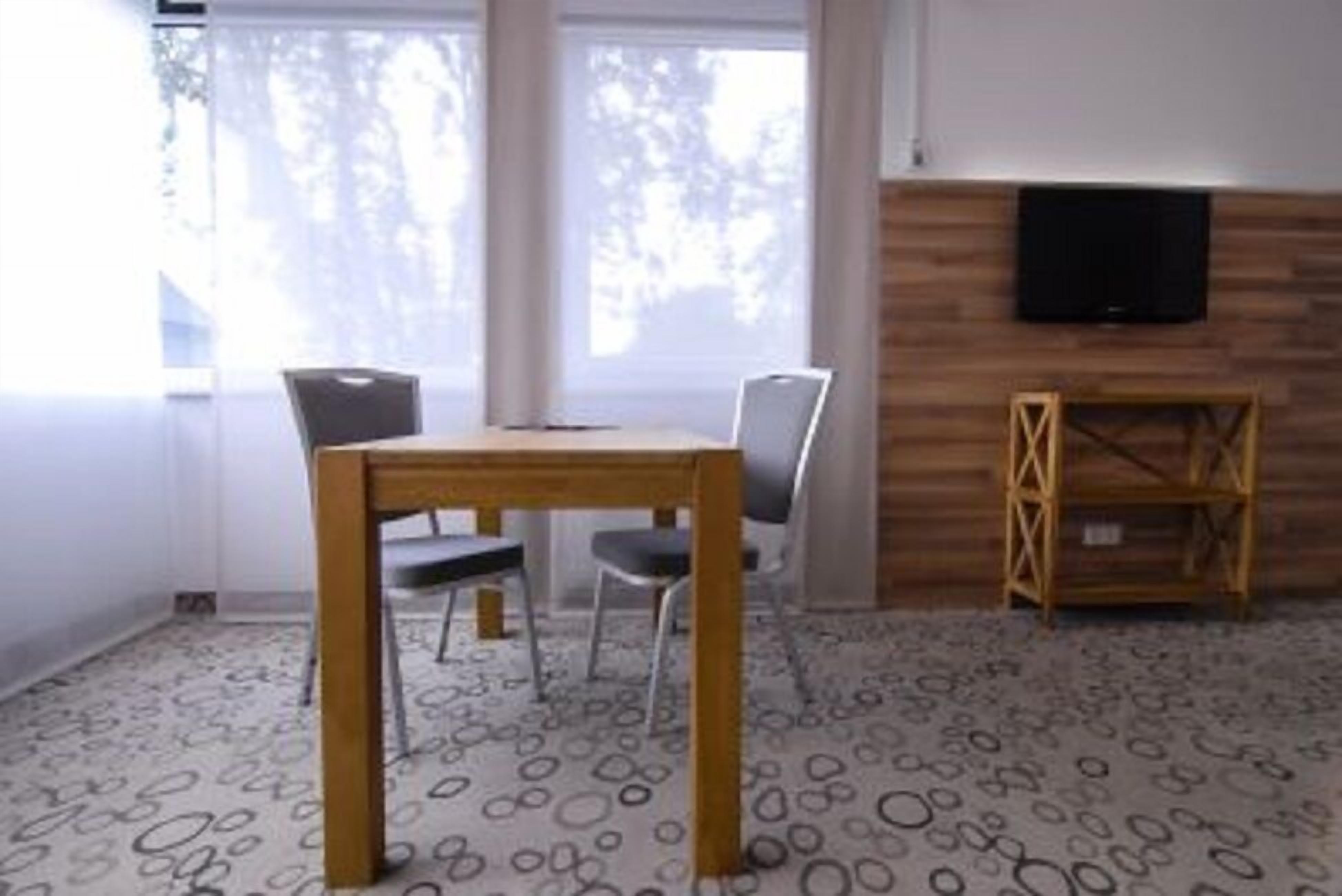 Huoneessa on puinen ruokapöytä ja -tuolit, seinällä televisio ja lattialla kuvioitu matto.