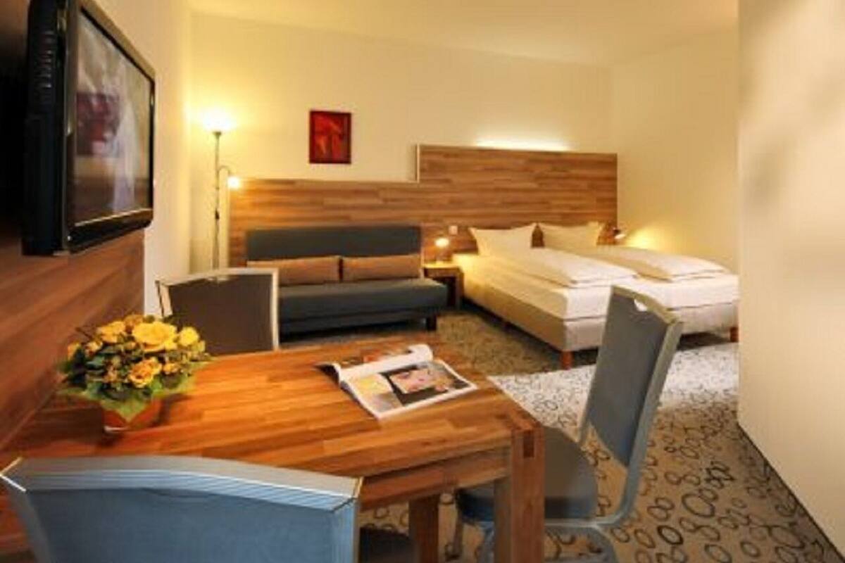 A hotel room with a wooden dining table, a sofa, a bed, and a TV mounted on the wall.