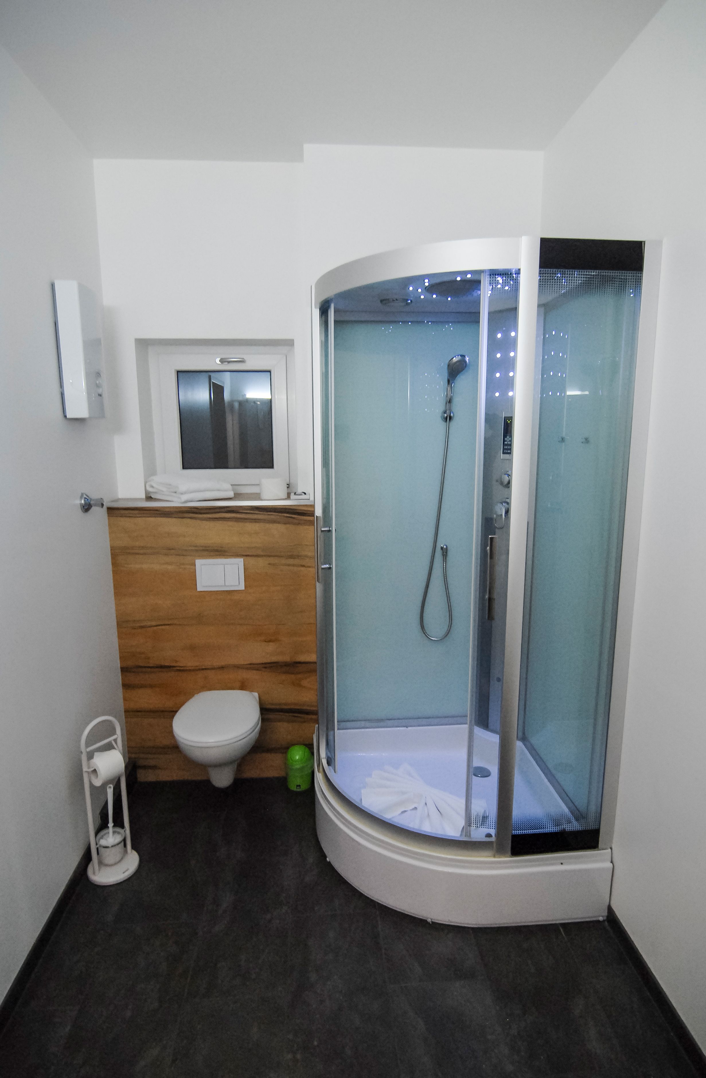 Un baño moderno con ducha envuelta en vidrio, tocador de madera, inodoro y un pequeño ventanal sobre el lavamanos.