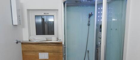 Un baño moderno con ducha envuelta en vidrio, tocador de madera, inodoro y un pequeño ventanal sobre el lavamanos.