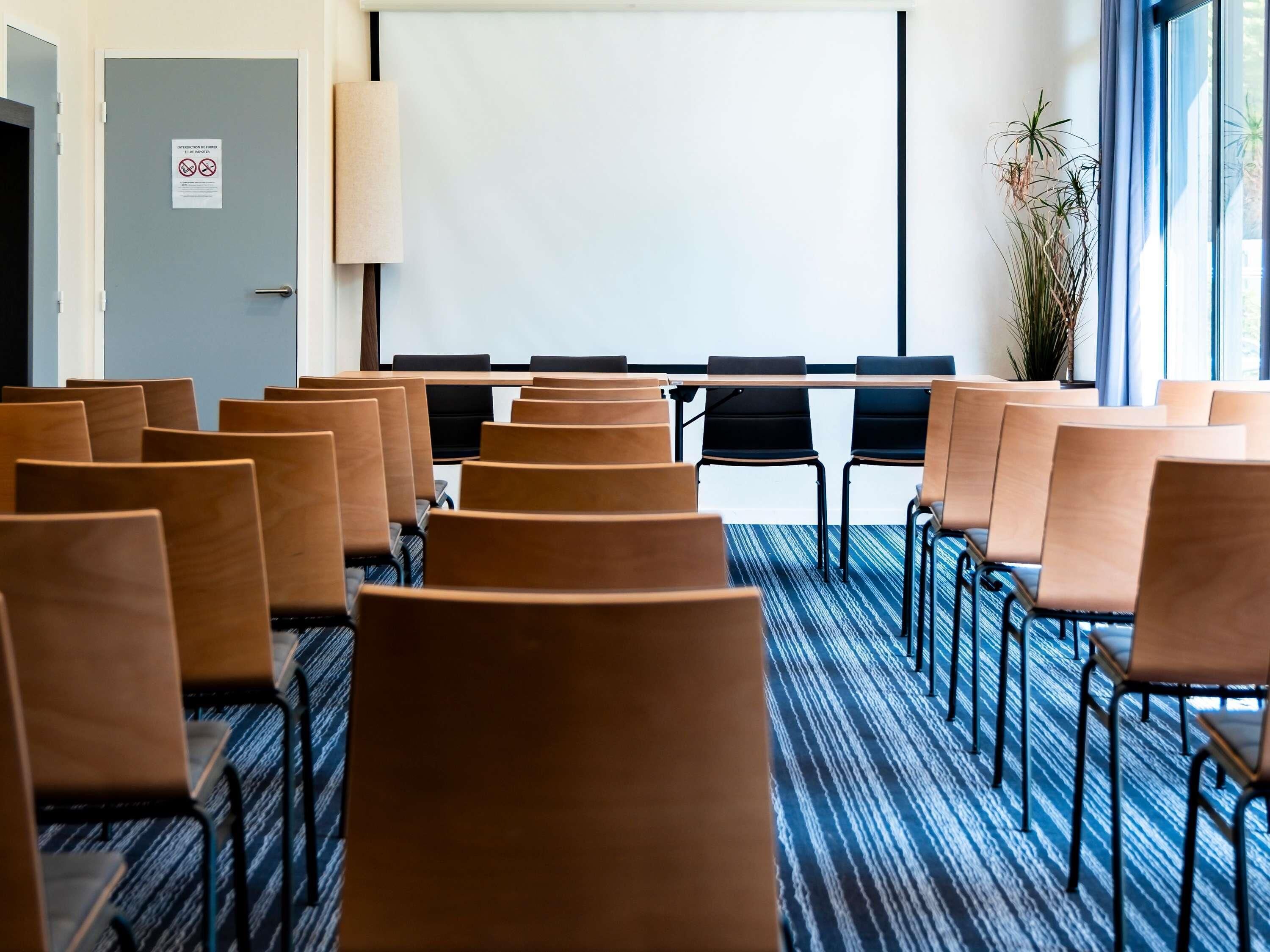 A conference room with rows of chairs, a projector screen, and a large window with a view of the outdoors.
