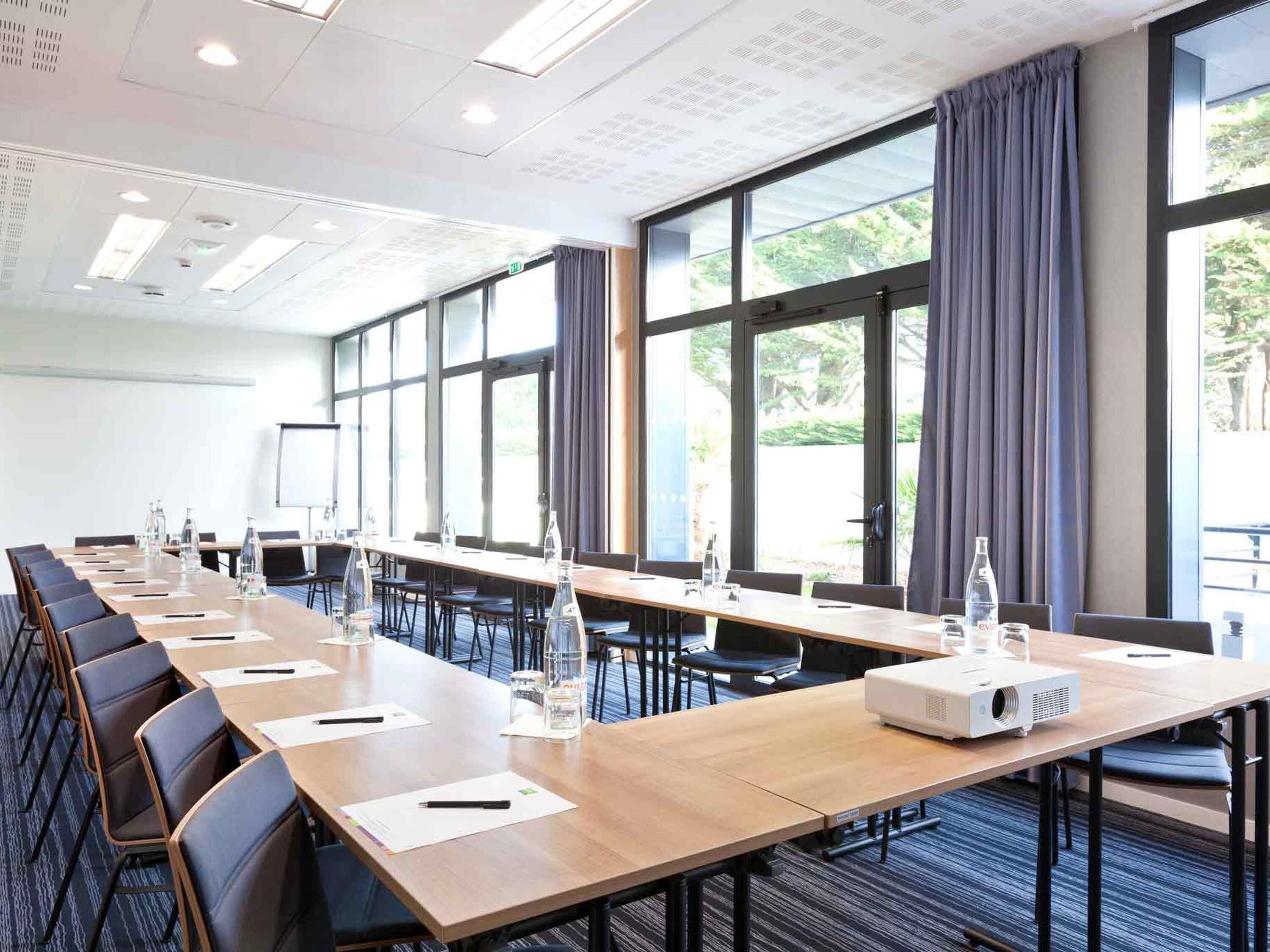A conference room with long tables, chairs, a projector, and large windows with curtains.