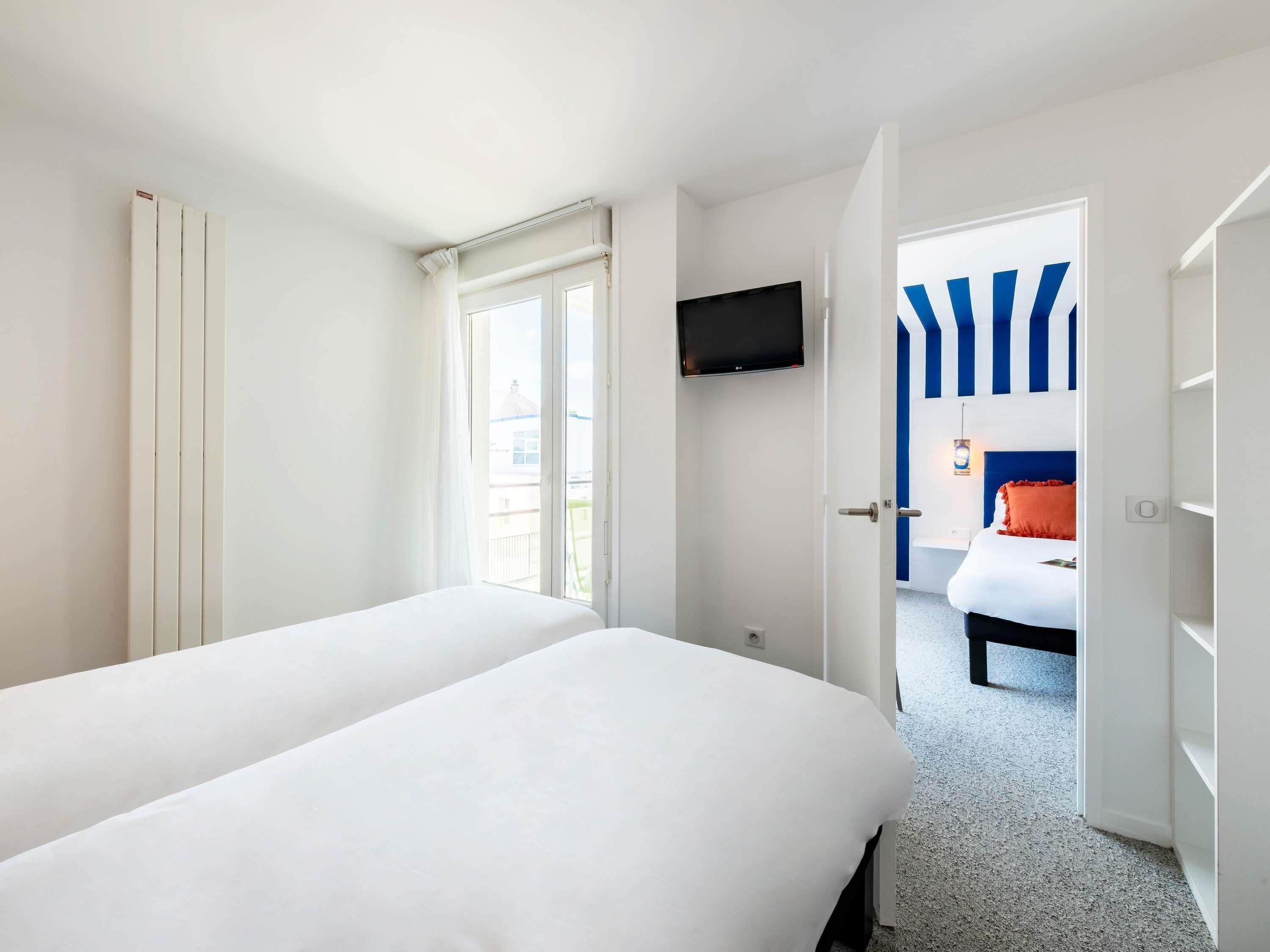 A hotel room with two beds, a TV mounted on the wall, and a view of another room with blue and white striped walls.